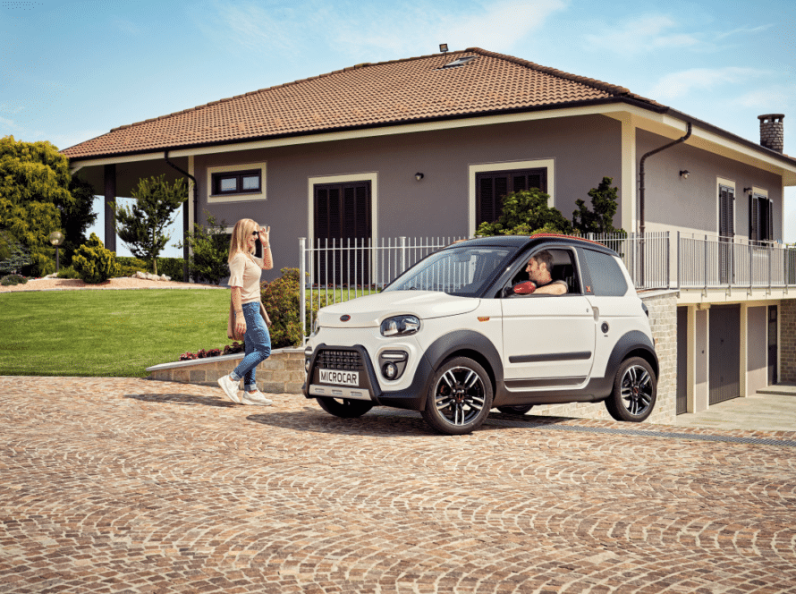Microcar blanco con un conductor al volante frente a una casa con jardín, un vehículo que necesita seguro coche sin carnet